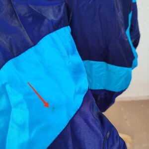 May include: A close-up of a vintage jacket in shades of blue and turquoise. The jacket features a colour-blocked design with a dark navy blue and turquoise colour scheme. The fabric appears to be a lightweight, possibly nylon, material.