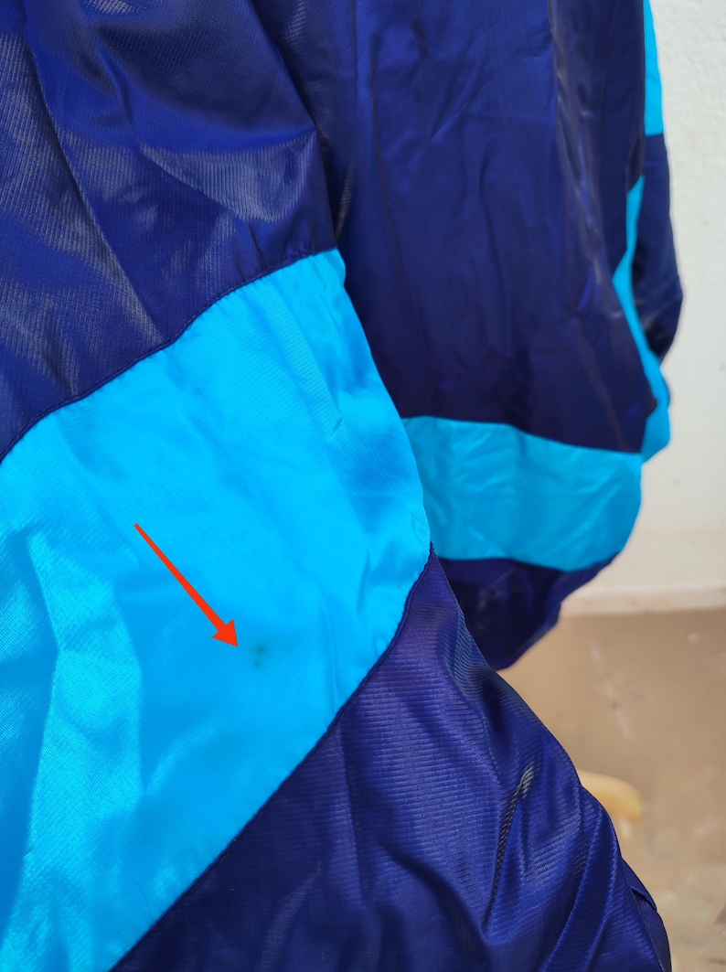 May include: A close-up of a vintage jacket in shades of blue and turquoise. The jacket features a colour-blocked design with a dark navy blue and turquoise colour scheme. The fabric appears to be a lightweight, possibly nylon, material.
