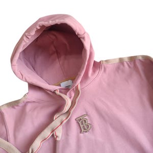 Puede incluir: Sudadera con capucha rosa claro con cord&oacute;n y capucha. La sudadera tiene un emblema plateado en el pecho. El cord&oacute;n es beige con una raya roja. La capucha est&aacute; forrada con una tela rosa m&aacute;s clara.