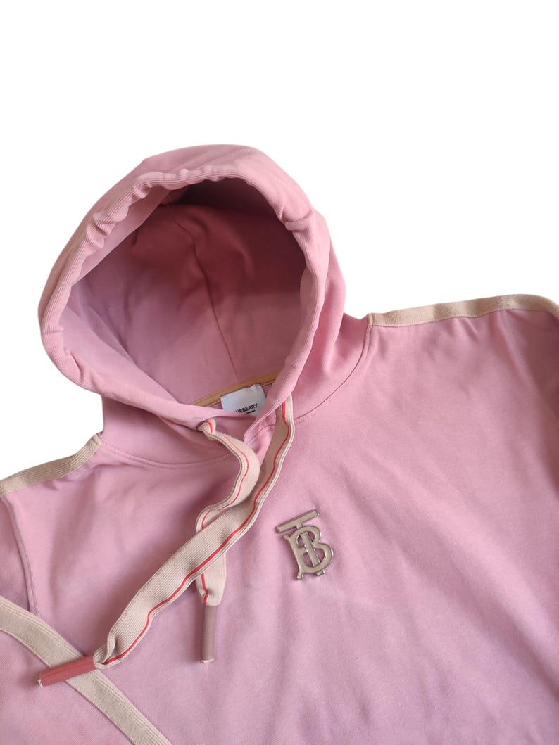 Puede incluir: Sudadera con capucha rosa claro con cord&oacute;n y capucha. La sudadera tiene un emblema plateado en el pecho. El cord&oacute;n es beige con una raya roja. La capucha est&aacute; forrada con una tela rosa m&aacute;s clara.