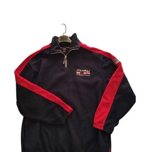 May include: A navy fleece pullover with red stripes on the sleeves. The top has a stand-up collar, a quarter-zip, and the "FUBU INTERNATIONAL" logo embroidered on the chest. A small flag patch is on the right sleeve.