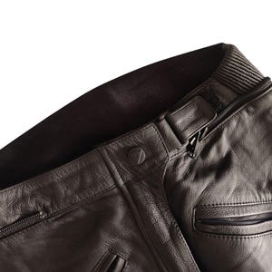 Może przedstawiać: Zbliżenie kołnierza czarnej sk&oacute;rzanej kurtki z prążkowanym dekoltem. Kurtka posiada zapięcie na zamek błyskawiczny, detal z guzikiem i małą kieszeń. Sk&oacute;ra ma teksturowany wygląd.