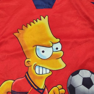 May include: Red soccer jersey featuring a cartoon character with yellow skin and spiky hair. The jersey has a blue collar with yellow and red stripes and a soccer ball graphic.