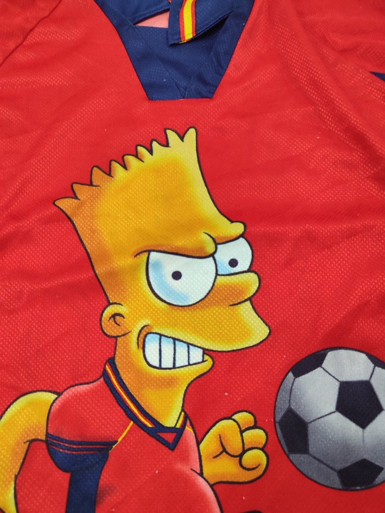 May include: Red soccer jersey featuring a cartoon character with yellow skin and spiky hair. The jersey has a blue collar with yellow and red stripes and a soccer ball graphic.