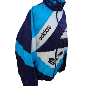 May include: A vintage Adidas track jacket in blue, white, and navy. The jacket features the Adidas logo and text in white. The design includes a high collar and a full-length zip. The jacket is made of a lightweight material.