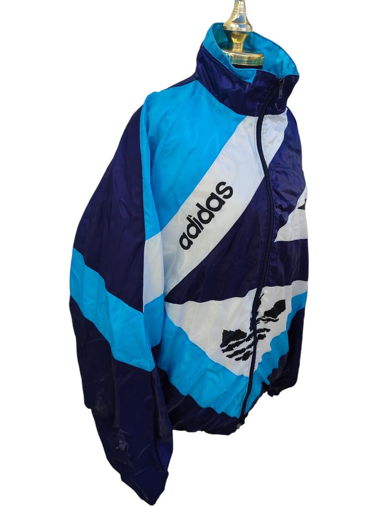 May include: A vintage Adidas track jacket in blue, white, and navy. The jacket features the Adidas logo and text in white. The design includes a high collar and a full-length zip. The jacket is made of a lightweight material.