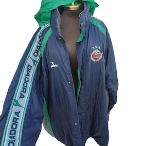 Könnte beinhalten: Marineblaue Kapuzenjacke mit grünen Akzenten und einer grünen Kapuze. Die Jacke trägt den Schriftzug "DIADORA" auf dem Ärmel und ein Logo auf der Brust. Die Jacke wird auf einer Schaufensterpuppe präsentiert.