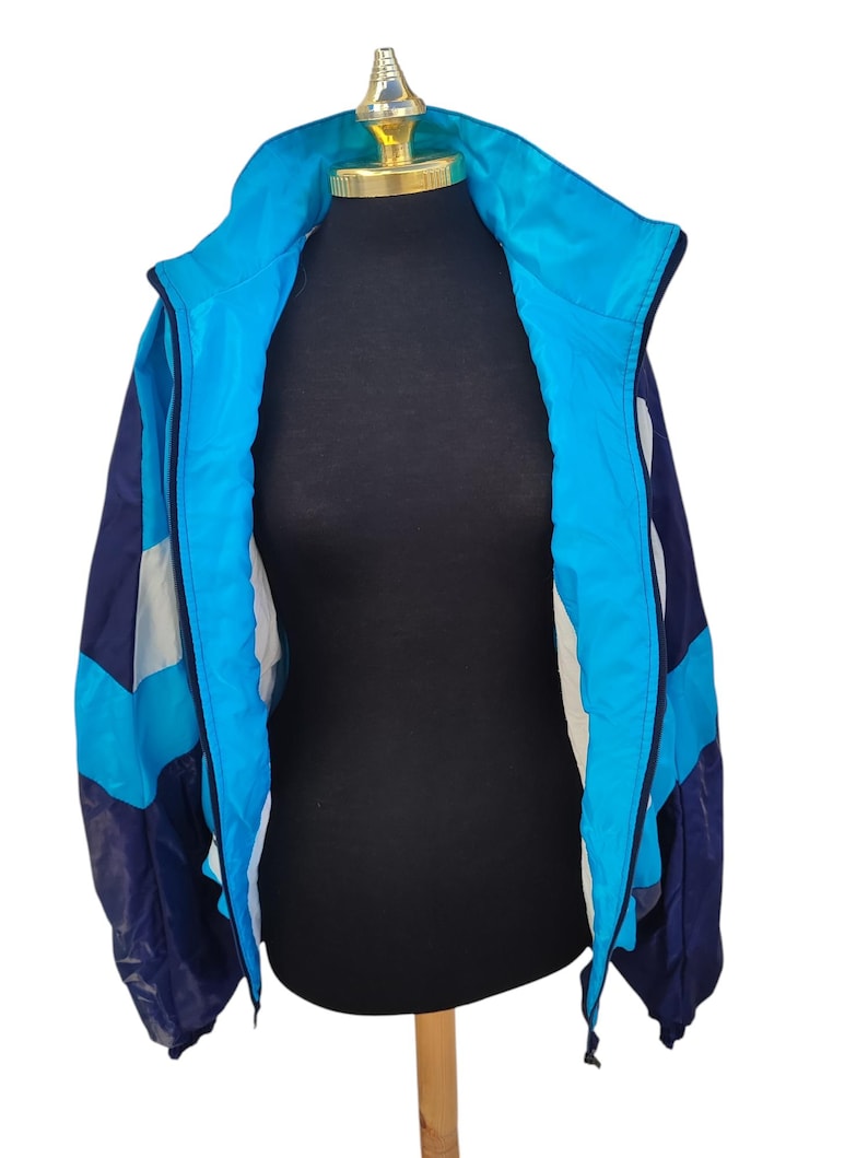 May include: A vintage track jacket in shades of blue, navy, and white, displayed on a mannequin. The jacket features a high collar, a full zip, and a colour-blocked design. The jacket is made of a lightweight material, perfect for layering.