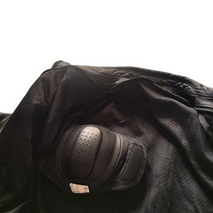 Może przedstawiać: Wnętrze czarnej kurtki motocyklowej z podszewką z siatki i ochraniaczem na łokieć. Ochraniacz na łokieć jest czarny, o zakrzywionym kształcie i małych otworach wentylacyjnych. Pasek na rzep zabezpiecza ochraniacz.