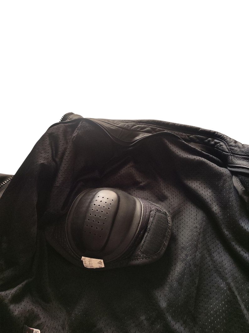 Może przedstawiać: Wnętrze czarnej kurtki motocyklowej z podszewką z siatki i ochraniaczem na łokieć. Ochraniacz na łokieć jest czarny, o zakrzywionym kształcie i małych otworach wentylacyjnych. Pasek na rzep zabezpiecza ochraniacz.