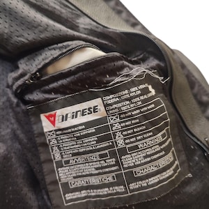 Może przedstawiać: Zbliżenie wnętrza czarnej sk&oacute;rzanej kurtki motocyklowej Dainese. Widoczna jest podszewka kurtki i kieszeń zapinana na zamek. Dołączona jest metka z instrukcjami pielęgnacji i ostrzeżeniami.