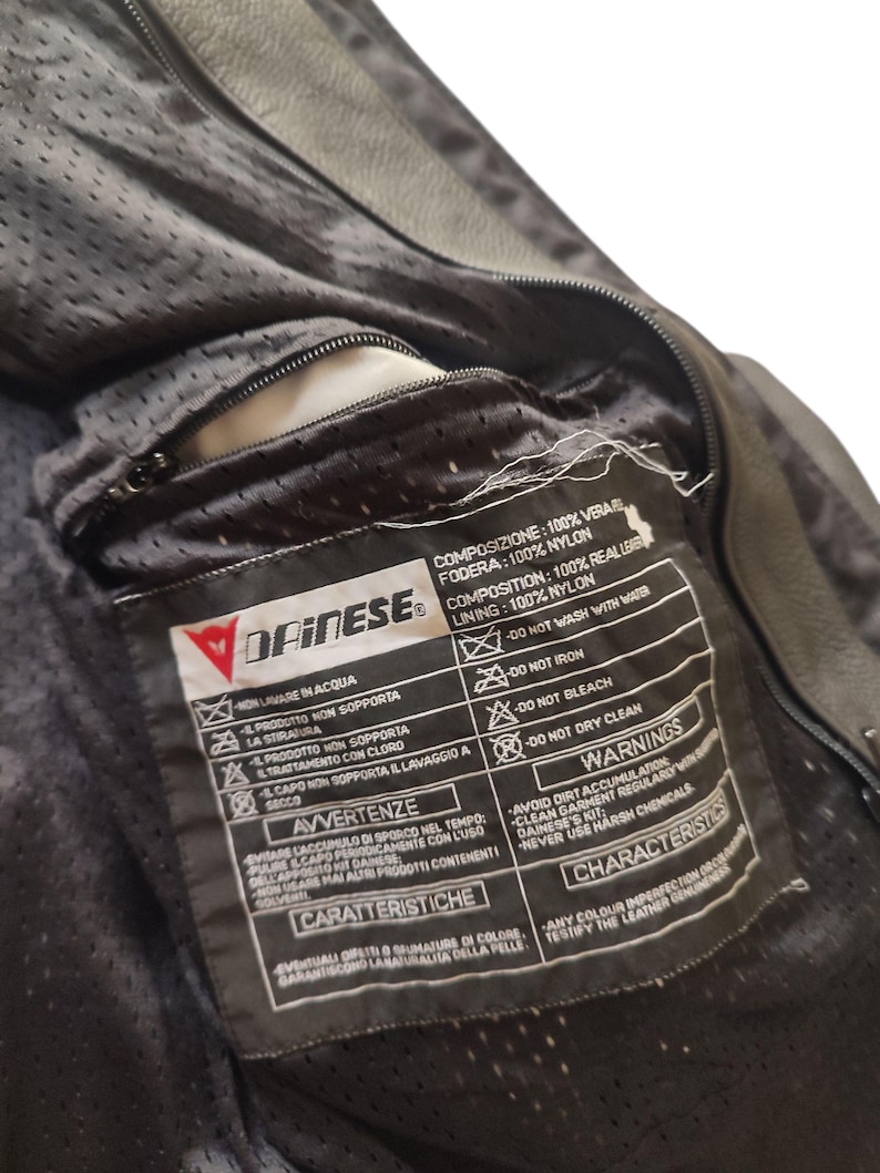 Może przedstawiać: Zbliżenie wnętrza czarnej sk&oacute;rzanej kurtki motocyklowej Dainese. Widoczna jest podszewka kurtki i kieszeń zapinana na zamek. Dołączona jest metka z instrukcjami pielęgnacji i ostrzeżeniami.