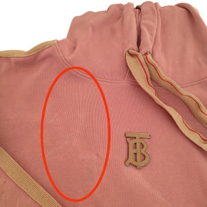 Puede incluir: Una sudadera con capucha rosa polvoriento con capucha con cord&oacute;n y un emblema met&aacute;lico dorado. La sudadera con capucha presenta un ribete beige a lo largo de la capucha y el cord&oacute;n, y un emblema met&aacute;lico dorado con las letras "TB".