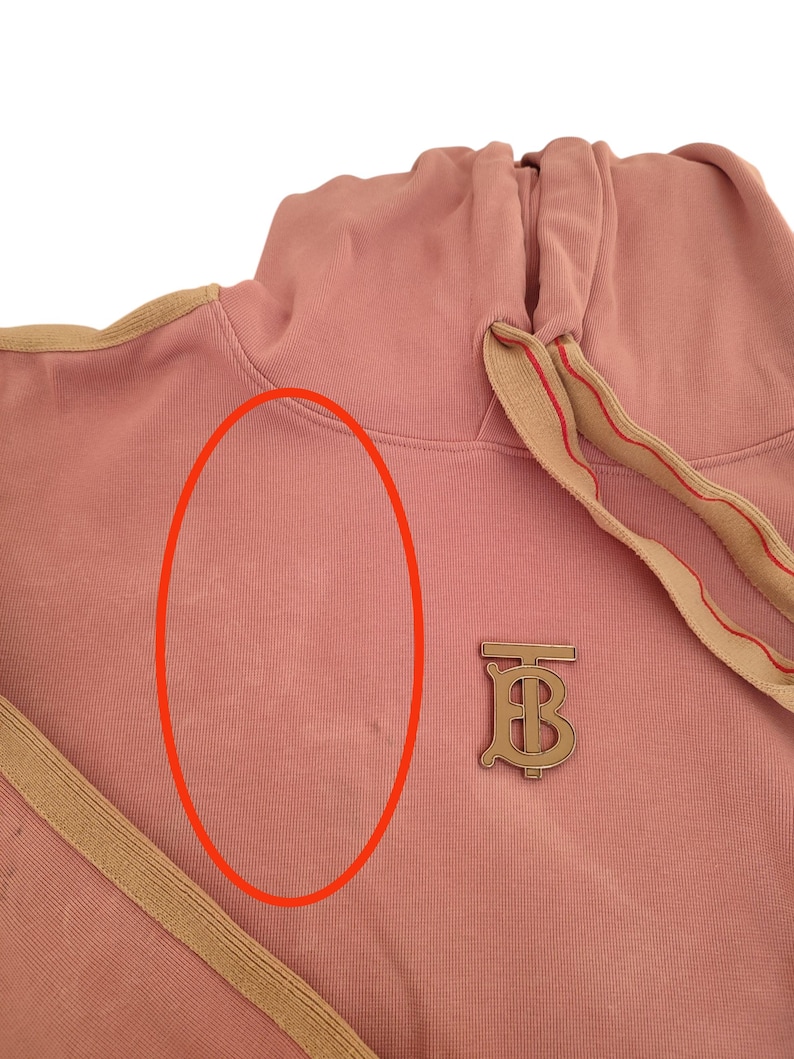 Puede incluir: Una sudadera con capucha rosa polvoriento con capucha con cord&oacute;n y un emblema met&aacute;lico dorado. La sudadera con capucha presenta un ribete beige a lo largo de la capucha y el cord&oacute;n, y un emblema met&aacute;lico dorado con las letras "TB".