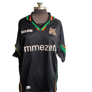 May include: A black short-sleeved soccer jersey featuring green and orange trim on the collar and sleeves. The shirt displays the Kelme logo, the text mmezeta, and the A.C. Venezia crest with a winged lion emblem on the chest area.