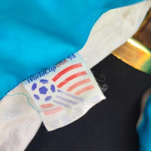 May include: A close-up of a World Cup USA 94 tag on a football jersey. The tag shows a football and the colours of the American flag. The jersey is a bright blue colour.