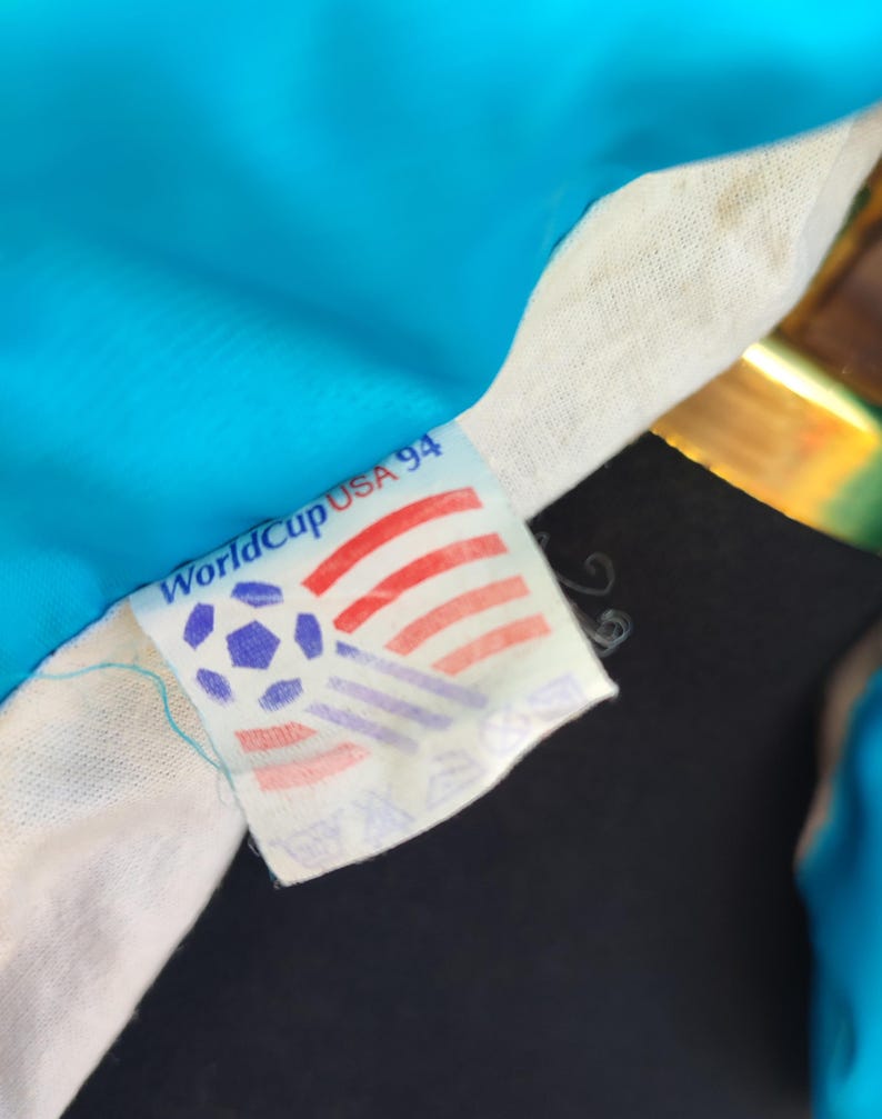 May include: A close-up of a World Cup USA 94 tag on a football jersey. The tag shows a football and the colours of the American flag. The jersey is a bright blue colour.