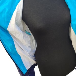 May include: A blue, white, and navy blue windbreaker jacket. The jacket has a hood and a zip closure. The interior lining is white. The jacket is displayed on a black mannequin.