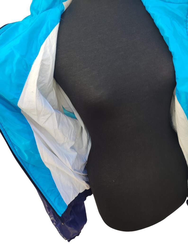 May include: A blue, white, and navy blue windbreaker jacket. The jacket has a hood and a zip closure. The interior lining is white. The jacket is displayed on a black mannequin.