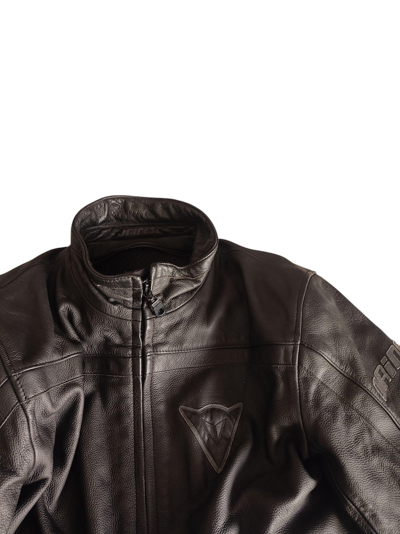 Może przedstawiać: Czarna sk&oacute;rzana kurtka motocyklowa ze st&oacute;jką i zapięciem na zamek błyskawiczny. Kurtka posiada tr&oacute;jkątne logo na piersi oraz nazwę marki na rękawie. Sk&oacute;ra ma teksturowany wygląd.