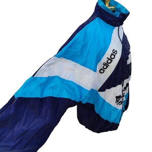 May include: A vintage Adidas track jacket in navy, white, and light blue. The jacket has a colour-block design with the Adidas logo on the chest and sleeve. It's made of a lightweight material and has a high collar.