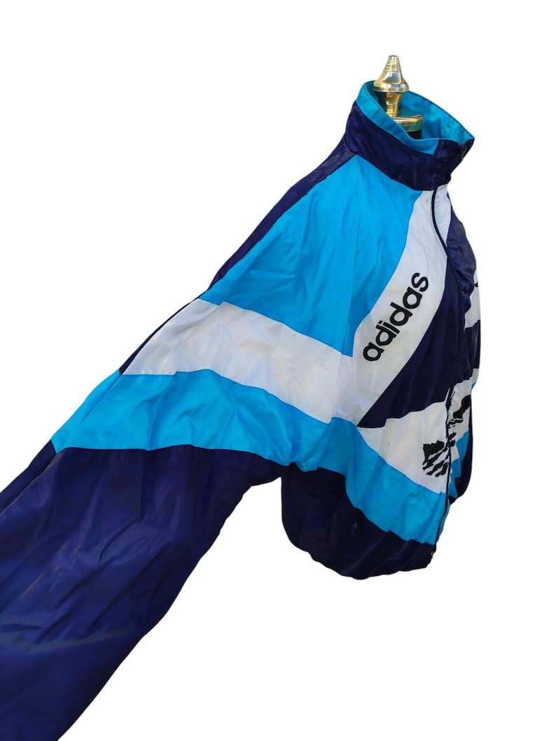 May include: A vintage Adidas track jacket in navy, white, and light blue. The jacket has a colour-block design with the Adidas logo on the chest and sleeve. It's made of a lightweight material and has a high collar.