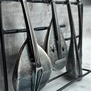 May include: A wall-mounted metal rack featuring three stylized, teardrop-shaped stringed instrument silhouettes. Each instrument is crafted from hammered silver-toned metal with vertical lines representing strings and a dark metal frame.