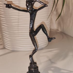 May include: A metal sculpture of a stylized figure in a dynamic pose, with one arm extended and the other raised to the head. The figure is dark gray and mounted on a base of stacked metal nuts. The sculpture is approximately 6 inches tall.