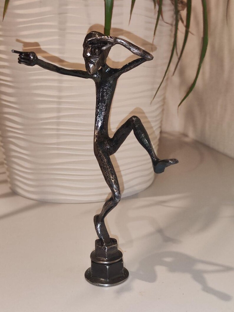 May include: A metal sculpture of a stylized figure in a dynamic pose, with one arm extended and the other raised to the head. The figure is dark gray and mounted on a base of stacked metal nuts. The sculpture is approximately 6 inches tall.