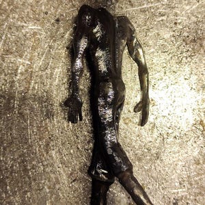 Pu&ograve; includere: Una statuetta metallica scura di una figura umanoide senza testa in posa di cammino. La scultura presenta una superficie testurizzata e riflettente con segni di saldatura visibili e una finitura color bronzo.