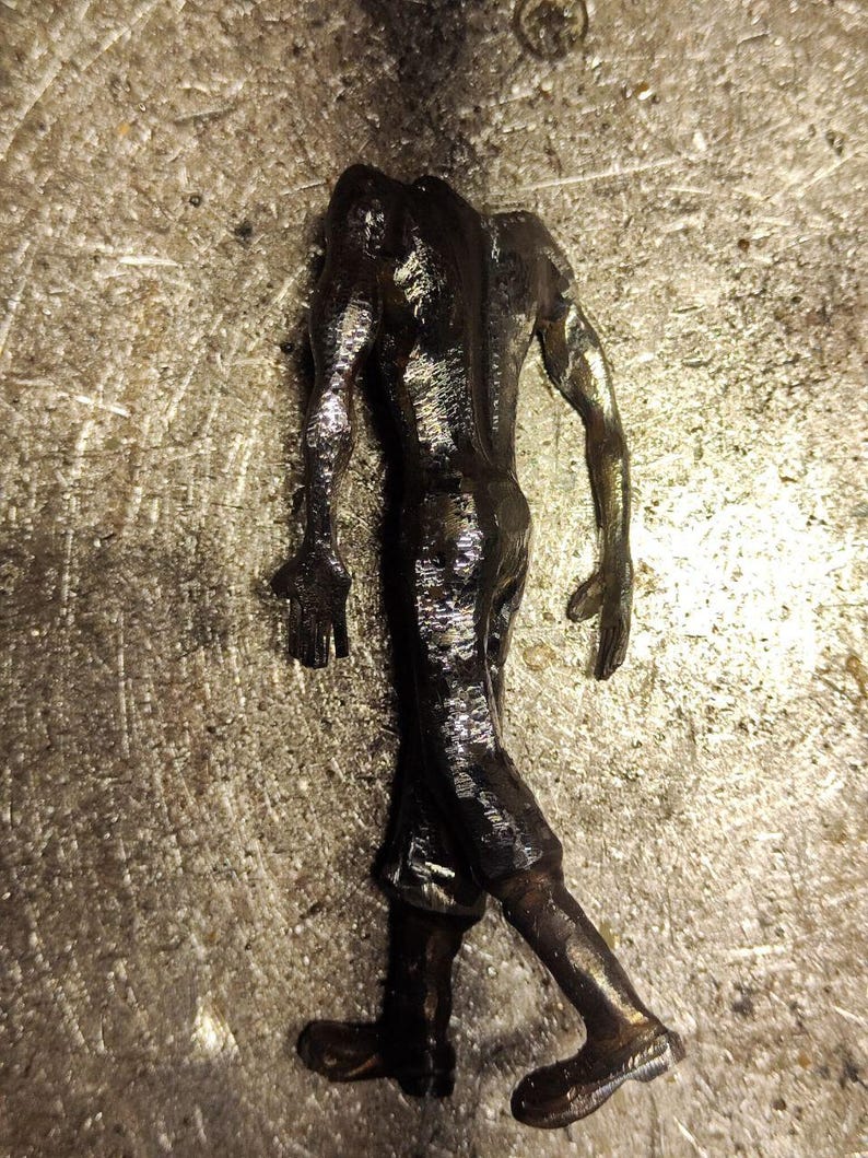 Pu&ograve; includere: Una statuetta metallica scura di una figura umanoide senza testa in posa di cammino. La scultura presenta una superficie testurizzata e riflettente con segni di saldatura visibili e una finitura color bronzo.