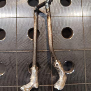 Pu&ograve; includere: Un paio di gambe in metallo realizzate con aste in acciaio scuro ossidato collegate in alto da una barra orizzontale. Le sezioni inferiori presentano piedi metallici testurizzati color argento saldati alle estremit&agrave; delle aste verticali.