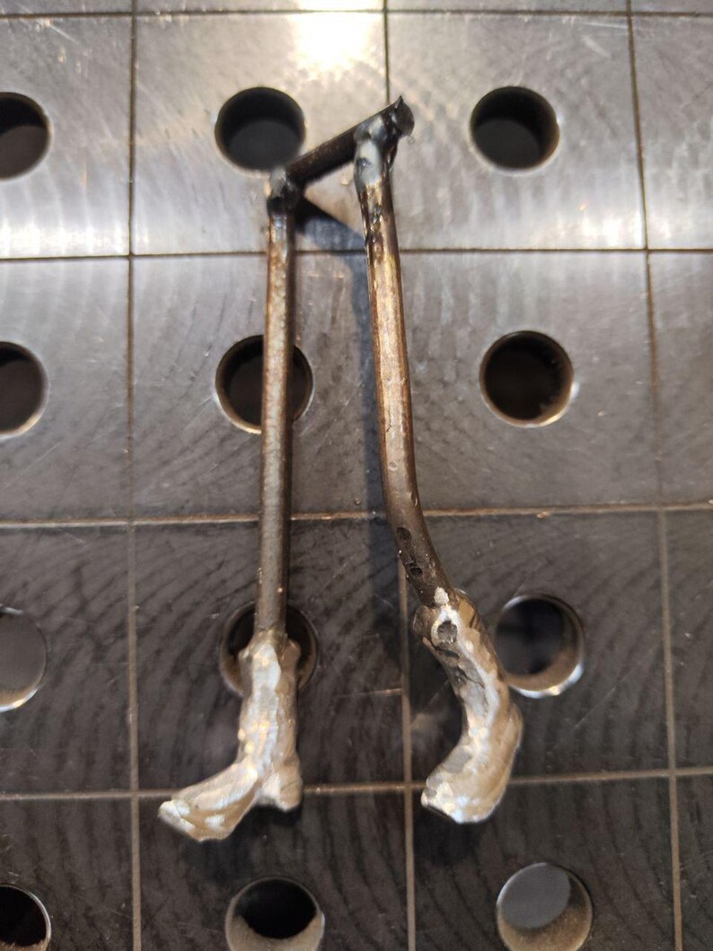 Pu&ograve; includere: Un paio di gambe in metallo realizzate con aste in acciaio scuro ossidato collegate in alto da una barra orizzontale. Le sezioni inferiori presentano piedi metallici testurizzati color argento saldati alle estremit&agrave; delle aste verticali.