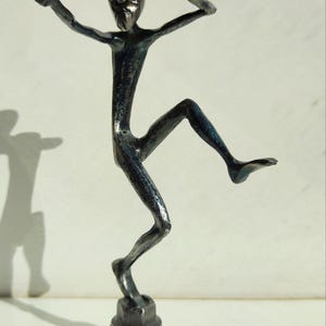 May include: A dark metal sculpture of a person in a dynamic pose, with one arm extended and the other hand shading the eyes. The figure is mounted on a hexagonal base, creating a striking contrast.