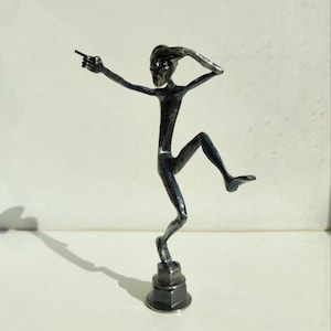 May include: A dark metal sculpture of a slender figure in a dynamic pose, with one arm outstretched and the other raised to the head. The figure is balanced on one leg, with the other leg bent. The sculpture is mounted on a hexagonal base.