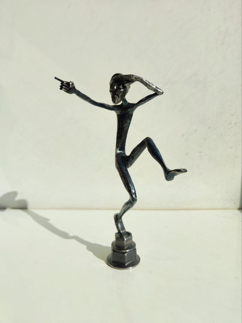 May include: A dark metal sculpture of a slender figure in a dynamic pose, with one arm outstretched and the other raised to the head. The figure is balanced on one leg, with the other leg bent. The sculpture is mounted on a hexagonal base.