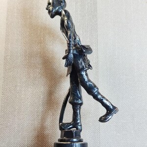 Pu&ograve; includere: Una statuetta metallica scura di una persona in posa di cammino, caratterizzata da una superficie ruvida, un piccolo chignon e una borsa in vita. La figura poggia su una base circolare ed &egrave; ritratta di profilo, mostrando una corporatura snella.