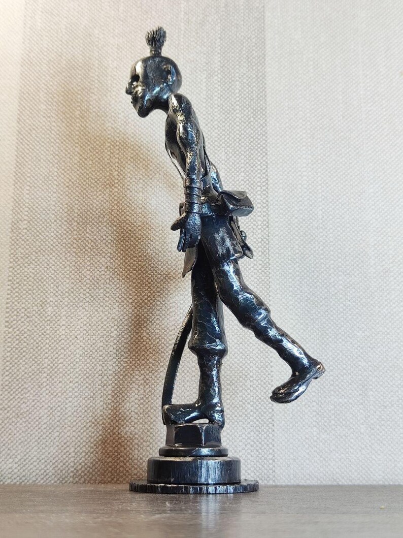 Pu&ograve; includere: Una statuetta metallica scura di una persona in posa di cammino, caratterizzata da una superficie ruvida, un piccolo chignon e una borsa in vita. La figura poggia su una base circolare ed &egrave; ritratta di profilo, mostrando una corporatura snella.