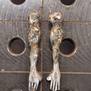 Pu&ograve; includere: Due sculture di braccia metalliche realizzate in acciaio saldato, caratterizzate da una superficie ruvida e testurizzata con cordoni di saldatura visibili e finiture color argento brunito. Ogni braccio presenta una spalla arrotondata, un bicipite definito e una mano con cinque dita.