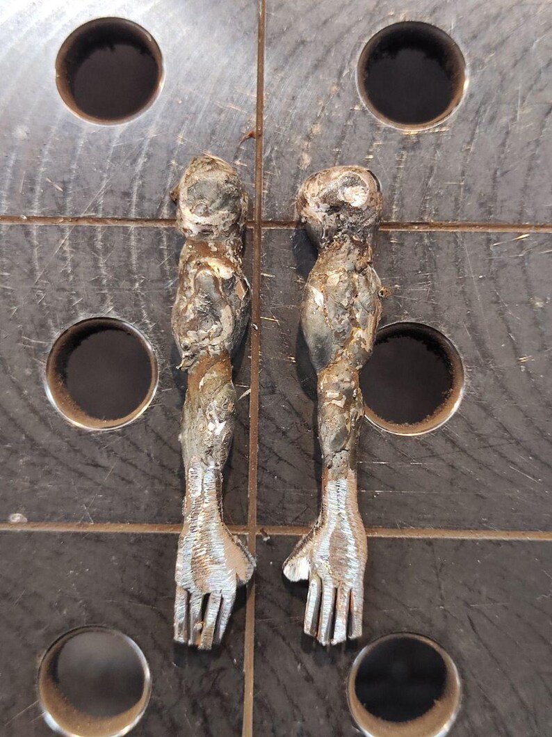 Pu&ograve; includere: Due sculture di braccia metalliche realizzate in acciaio saldato, caratterizzate da una superficie ruvida e testurizzata con cordoni di saldatura visibili e finiture color argento brunito. Ogni braccio presenta una spalla arrotondata, un bicipite definito e una mano con cinque dita.