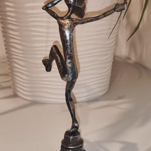 May include: A metal sculpture of a figure in a dancing pose. The sculpture is made of silver-colored metal and is mounted on a base of stacked nuts and bolts. The figure has one leg bent and arms outstretched.