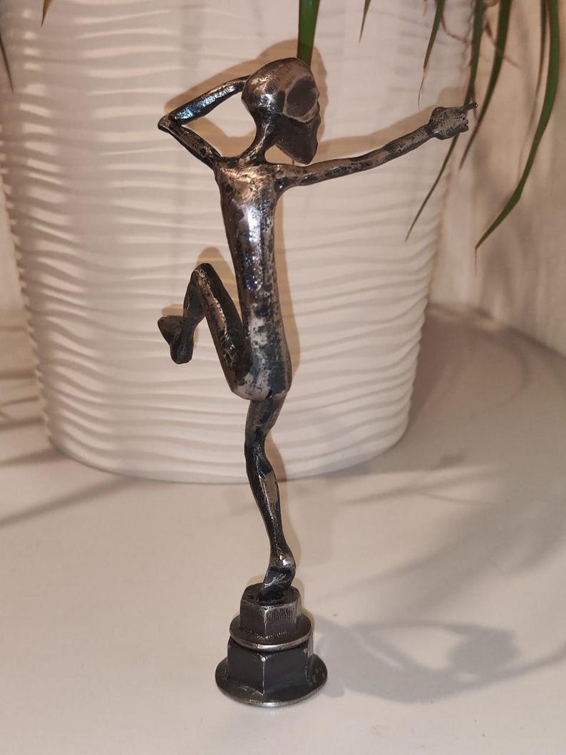 May include: A metal sculpture of a figure in a dancing pose. The sculpture is made of silver-colored metal and is mounted on a base of stacked nuts and bolts. The figure has one leg bent and arms outstretched.