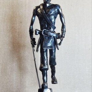 Pu&ograve; includere: Una scultura metallica dai toni scuri raffigurante una figura in piedi con un'acconciatura a chignon, che indossa una cintura con fibbia e una fascia sul petto. La figura tiene una spada sottile e dritta nella mano destra su una base circolare.
