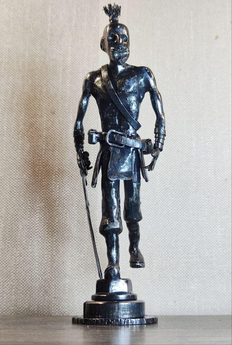Pu&ograve; includere: Una scultura metallica dai toni scuri raffigurante una figura in piedi con un'acconciatura a chignon, che indossa una cintura con fibbia e una fascia sul petto. La figura tiene una spada sottile e dritta nella mano destra su una base circolare.