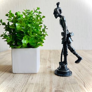 Pu&ograve; includere: Una statuetta metallica scura di una persona curva con un'acconciatura a chignon, in piedi su una base circolare. La figura &egrave; raffigurata in una postura inclinata in avanti con una gamba sollevata, indossando abiti semplici e una piccola borsa.