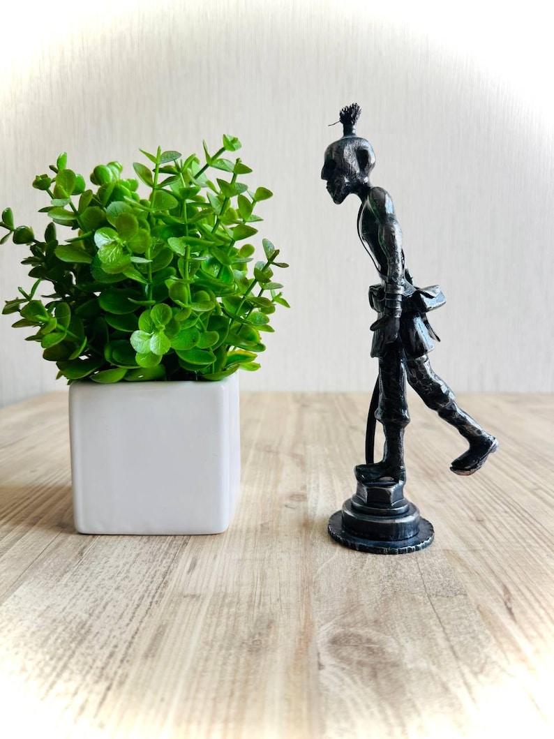 Pu&ograve; includere: Una statuetta metallica scura di una persona curva con un'acconciatura a chignon, in piedi su una base circolare. La figura &egrave; raffigurata in una postura inclinata in avanti con una gamba sollevata, indossando abiti semplici e una piccola borsa.