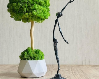 May include: A decorative topiary tree with a bright green moss canopy, a light-colored trunk, and a white geometric concrete pot, positioned next to a slender, dark metal sculpture of a figure in a graceful, reaching pose on a circular base.