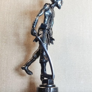Pu&ograve; includere: Una scultura metallica dai toni scuri raffigurante una figura umanoide stilizzata in piedi su una base circolare. Il personaggio ha un'acconciatura a punta, un fisico snello ed &egrave; raffigurato in posa di cammino mentre impugna una lama curva.