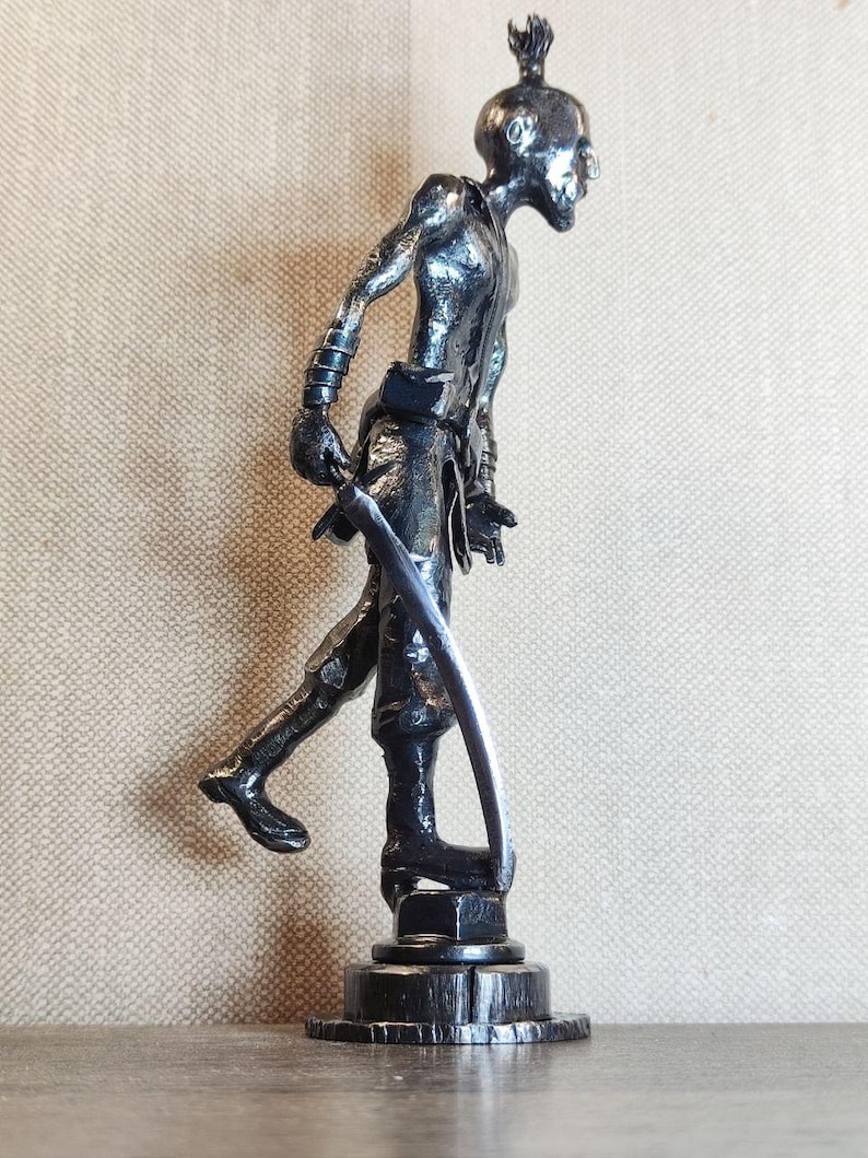 Pu&ograve; includere: Una scultura metallica dai toni scuri raffigurante una figura umanoide stilizzata in piedi su una base circolare. Il personaggio ha un'acconciatura a punta, un fisico snello ed &egrave; raffigurato in posa di cammino mentre impugna una lama curva.