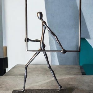 May include: A dark metallic sculpture of a stylized human figure in a walking pose, holding a large rectangular frame. The figure is attached to a rectangular base, and the entire piece features a textured, dark bronze-toned finish throughout.